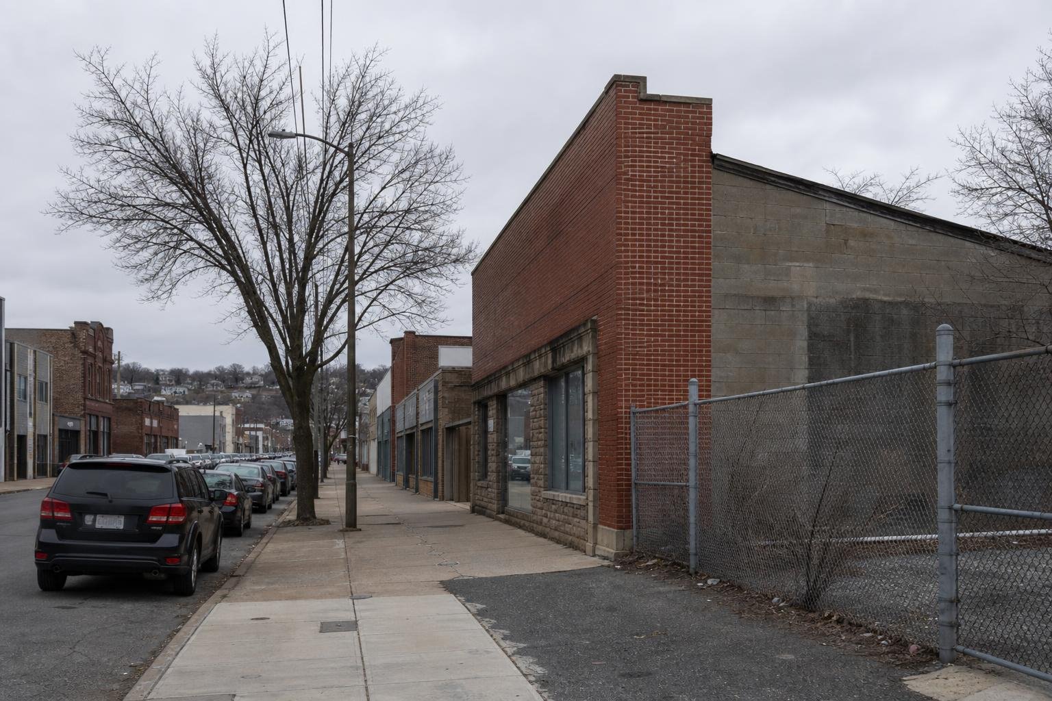 A quiet urban street with parked cars beside low-rise brick and concrete buildings under an overcast sky.