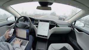 A person is pictured behind the wheel of an autonomous vehicle. The interior is gray and black with a large touch screen in the dash.